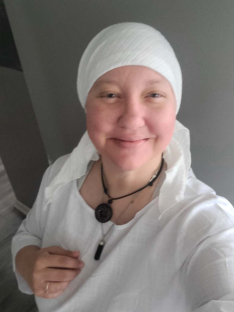 A woman smiling, wearing a white headscarf and a white tunic, holding her hands gently against her chest, standing in a softly lit room with a gray wall in the background.