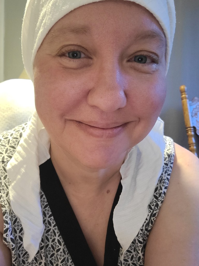 A smiling person wearing a white head wrap and a patterned top, sitting indoors with a warm expression.