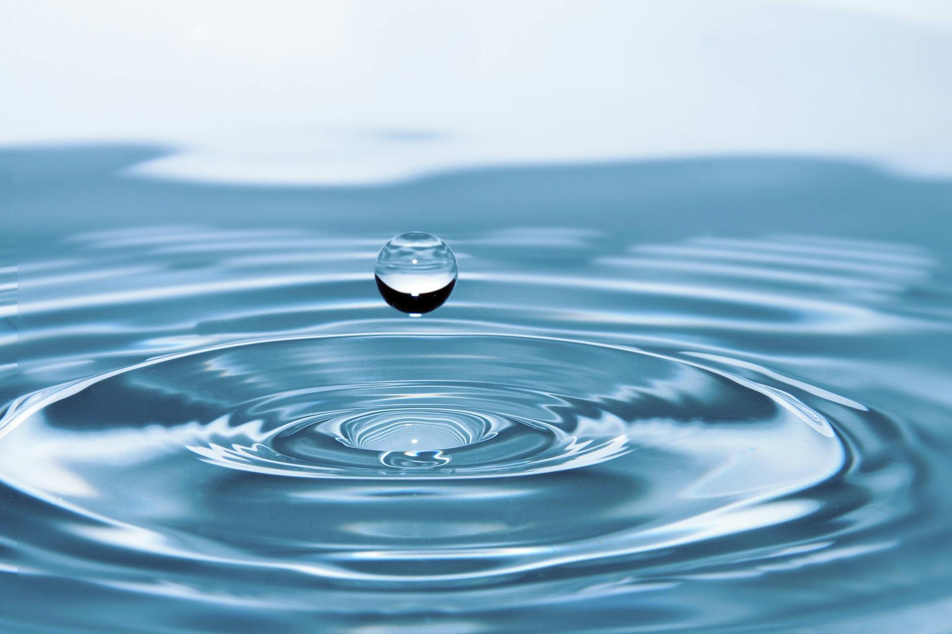 A close-up image of a water droplet falling onto the surface of water, creating ripples in a calm blue setting.