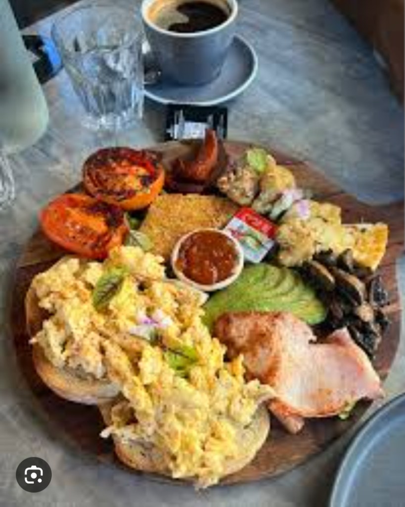 A wooden platter with a hearty breakfast spread including scrambled eggs on toast, grilled tomatoes, avocado, bacon, mushrooms, and a small dish of sauce, accompanied by a cup of coffee and a glass of water.